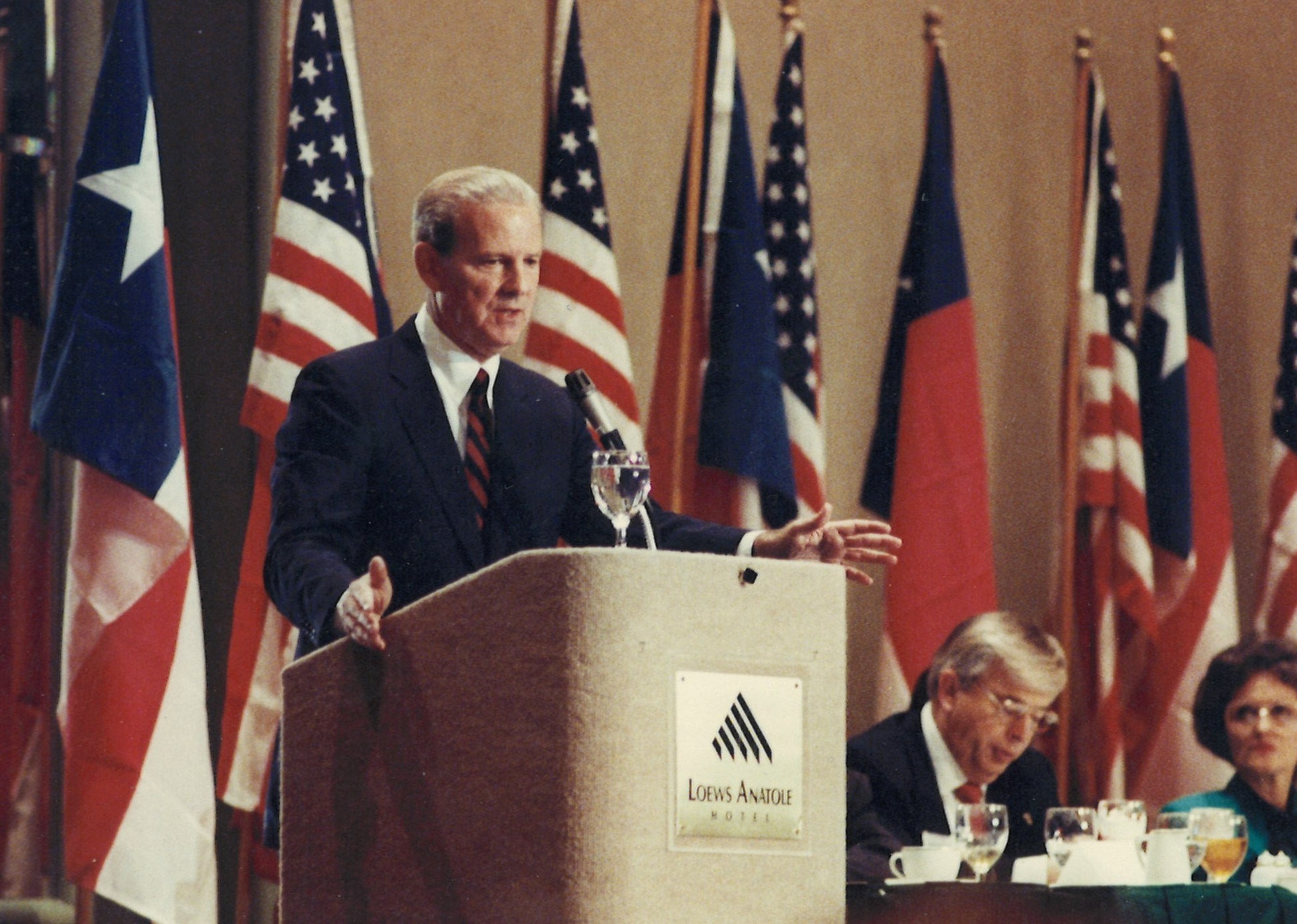 Former United States Secretary of State James Baker at a podium with the flags of the United States of America and the State of Texas behind him